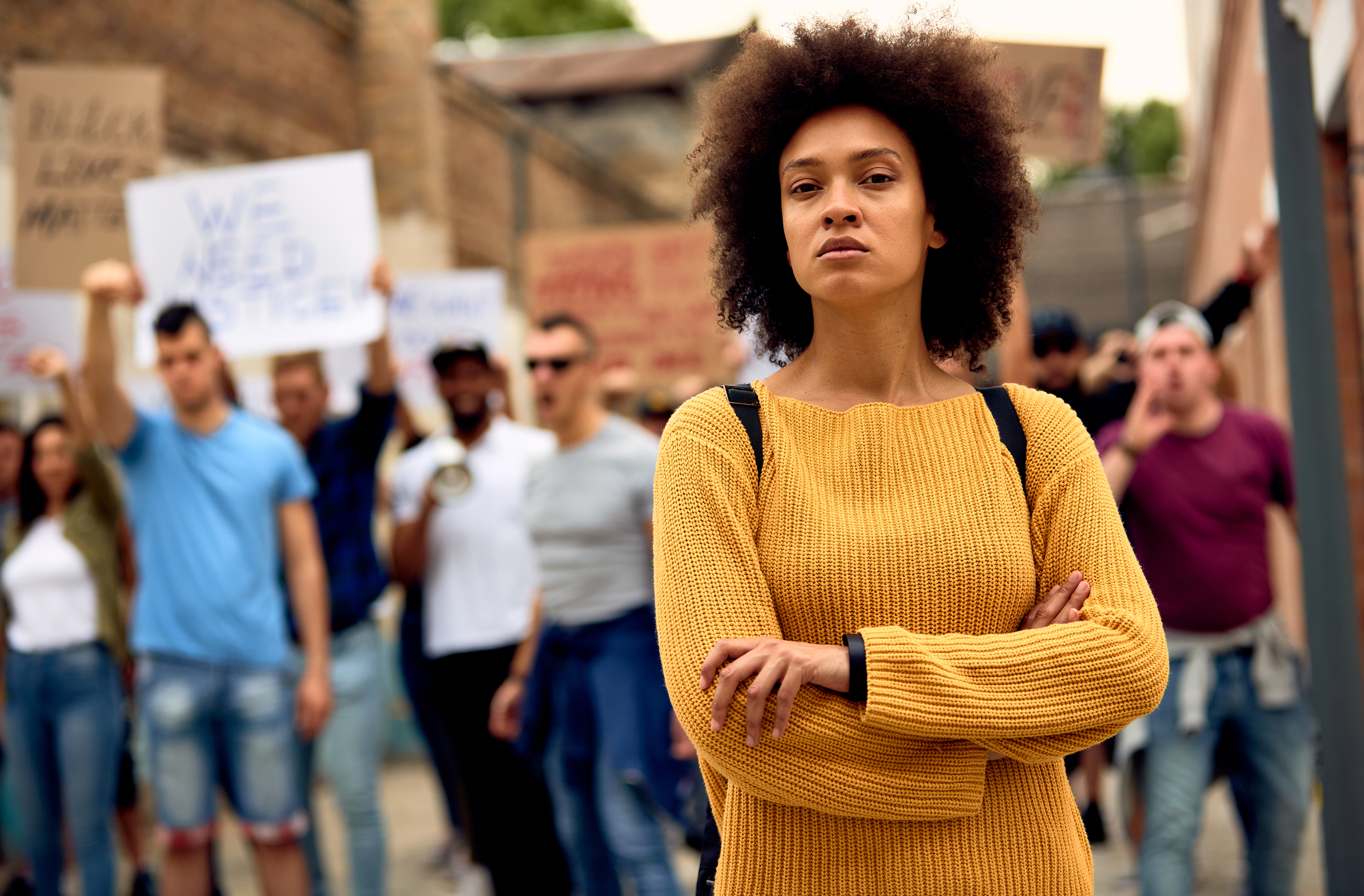 Young black woman with arms crossed standing in front of crowd of people on anti-racism protest.